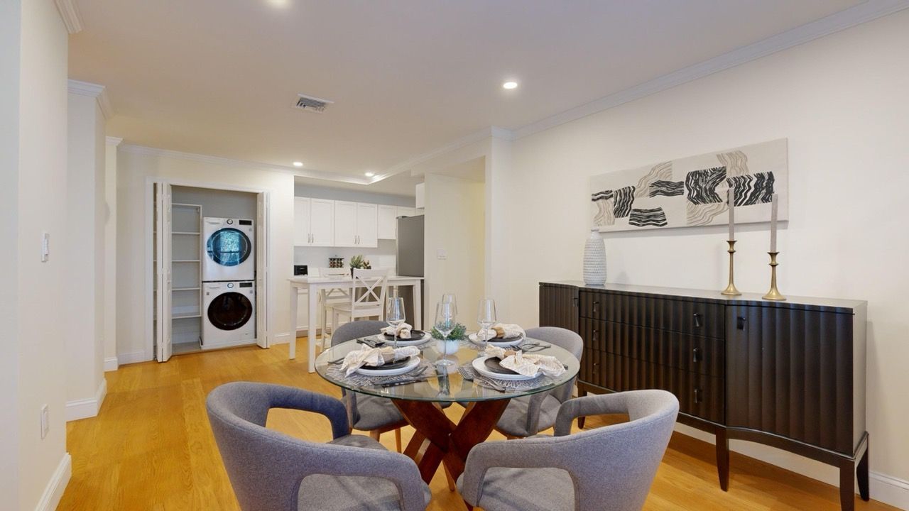 Dining area of a modern apartment with a round glass table, gray upholstered chairs, and a kitchen in view.
