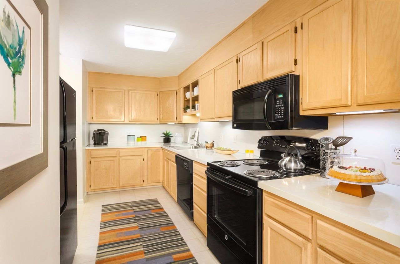 Kitchen in an apartment with light wood cabinets, black appliances, and a striped rug.