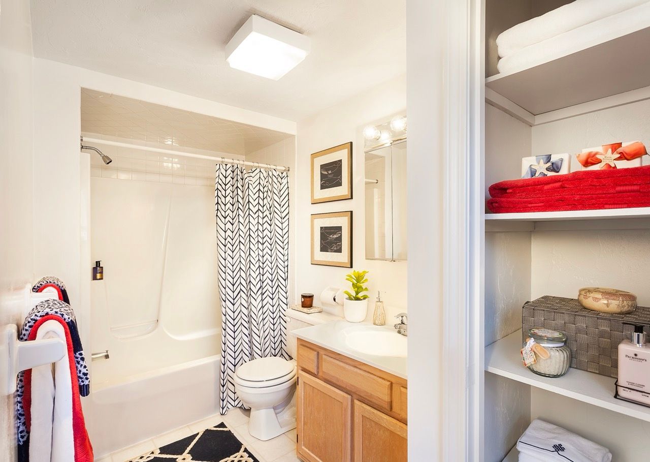 Bathroom in apartment with tub/shower, toilet, vanity, and open linen shelves.