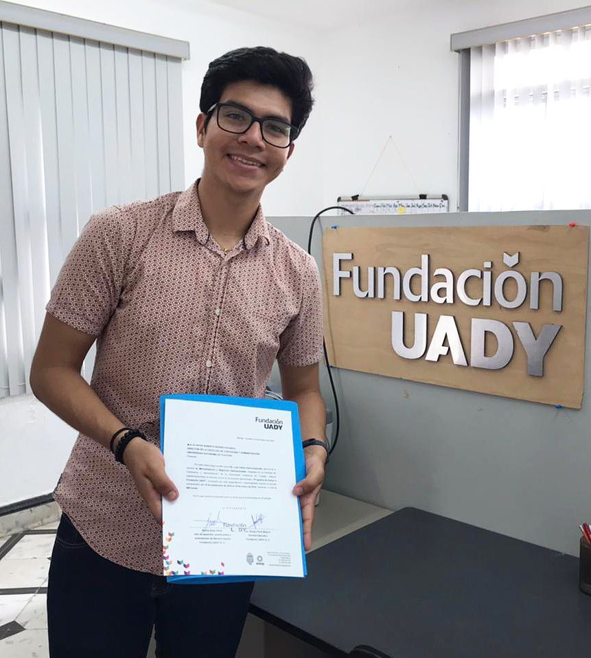 Un joven sostiene un certificado frente a un cartel que dice fundación uady