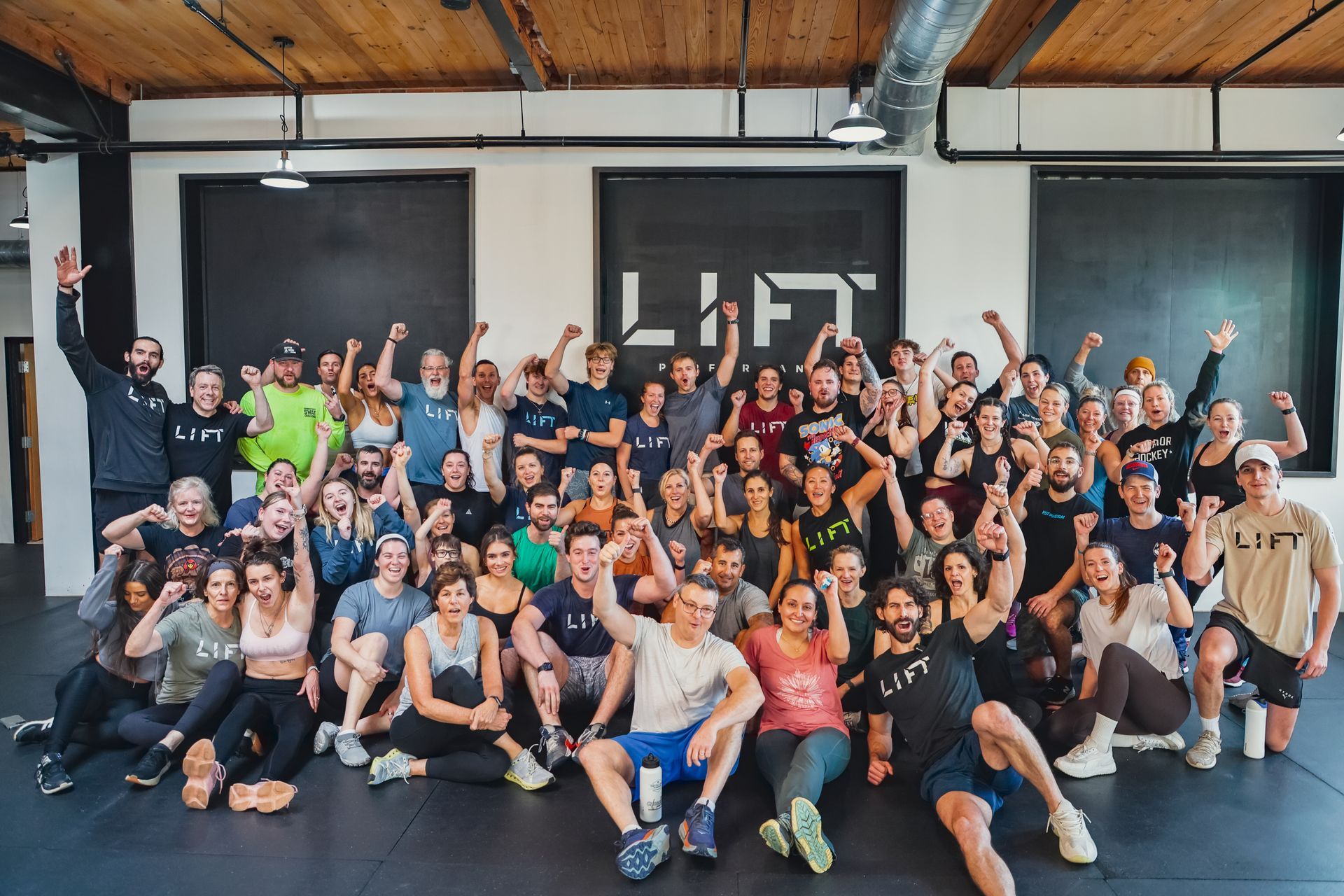 Large group of people, likely gym members, posing with raised arms in front of "LIFT" sign.