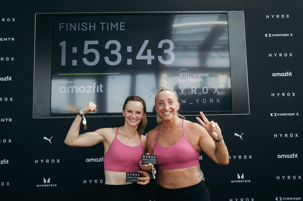 Two women in pink sports bras flexing before a finish time display reading 1:53:43