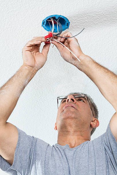 Electrician wiring a ceiling light fixture. Blue box, gray and white wires, focused expression.