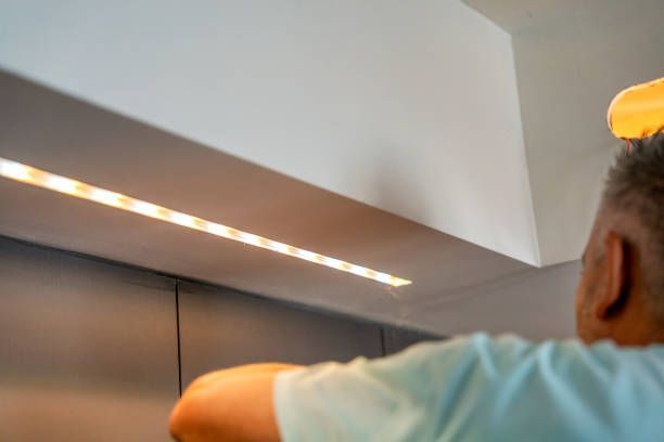 A person installing a warm LED strip light under a white cabinet.