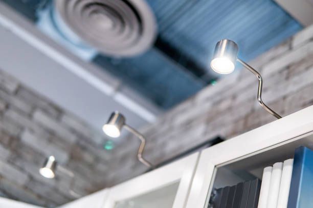 Three small, silver-toned spotlights illuminate books on a white shelf, near a brick wall and ceiling vent.