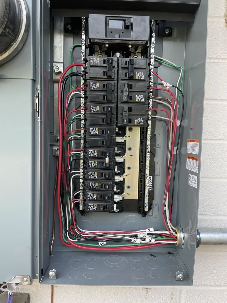 Electrical panel with numerous circuit breakers and wires. Gray metal box mounted on a wall.