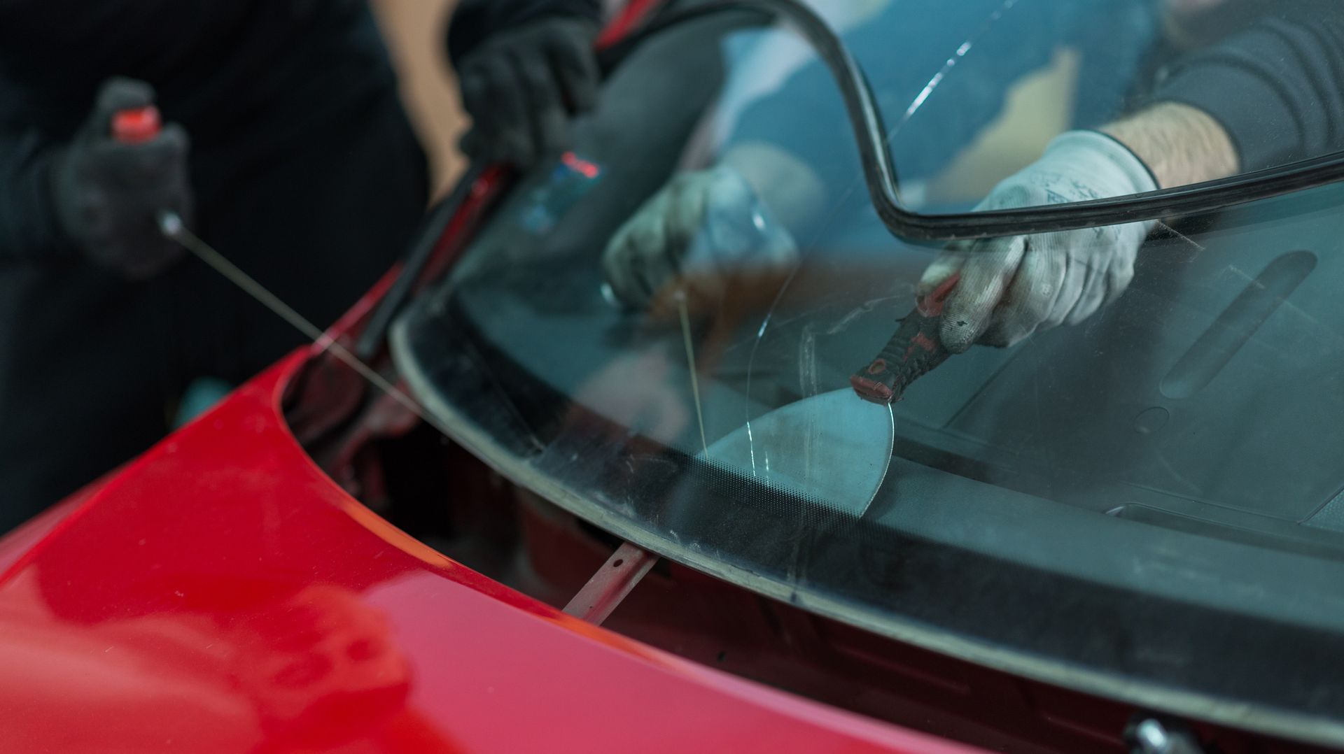 Close-up of a mechanic removing a damaged windshield off a red car’s front.