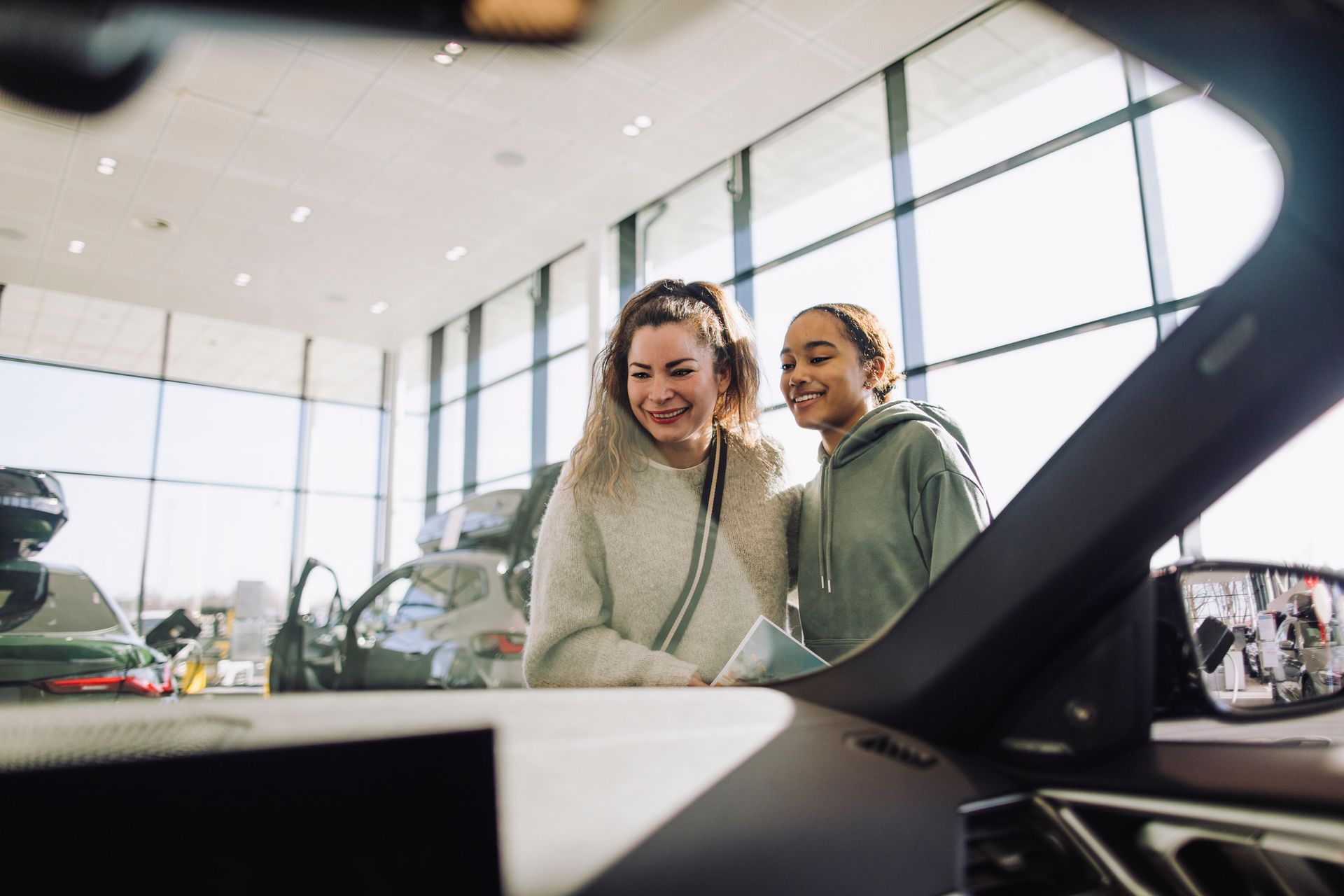 Excited mother and daughter by car in showroom seen through windshield. Excited mother and daughter by car in showroom seen through windshield.