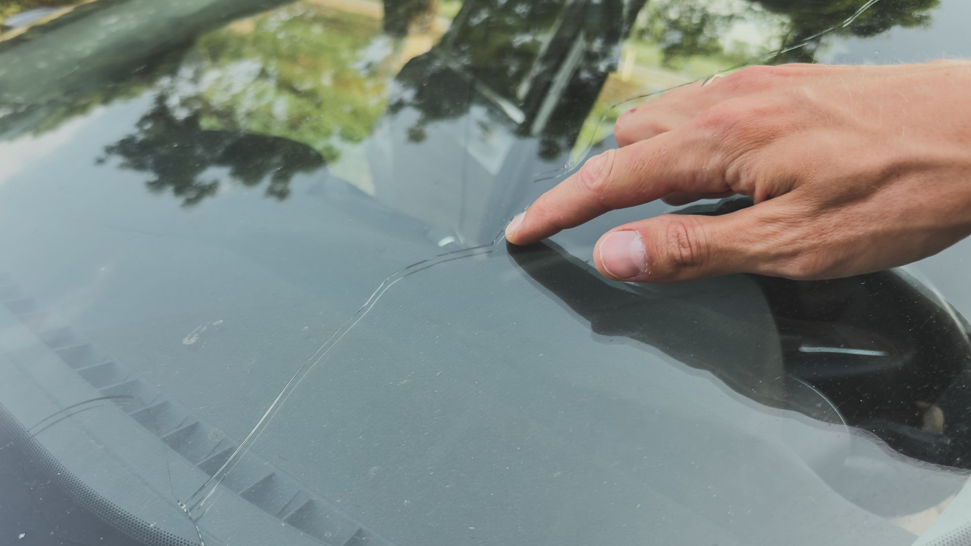 A hand pointing at a long crack in a car windshield.