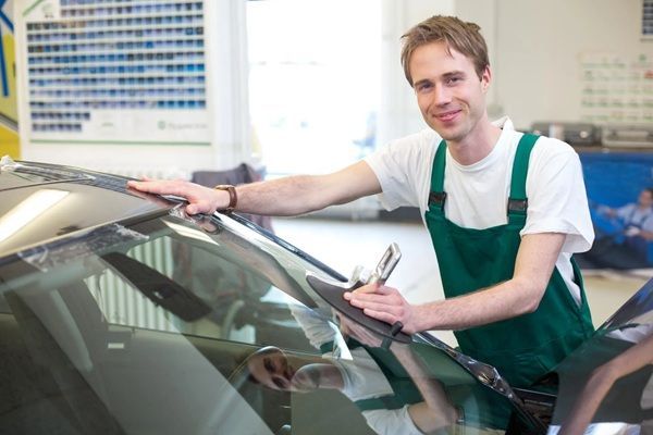 A man in green overalls is working on a car windshield.