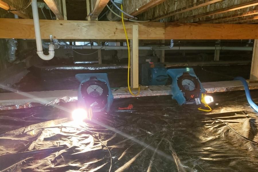 Two industrial fans drying a crawlspace with black plastic sheeting, lit by work lights.