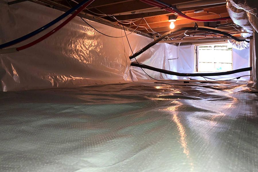Crawl space with white plastic sheeting on walls and floor, utility lines, and a distant window.