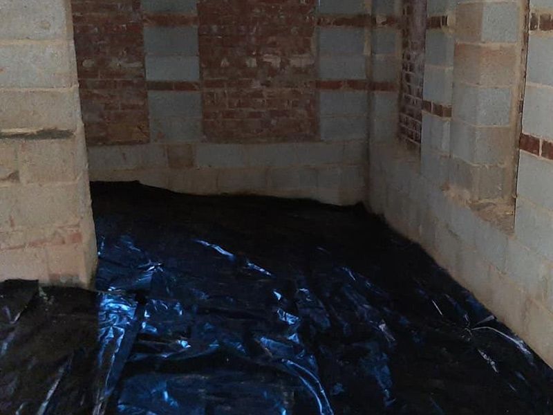 Black plastic sheeting covers the floor of a brick and cinder block basement.
