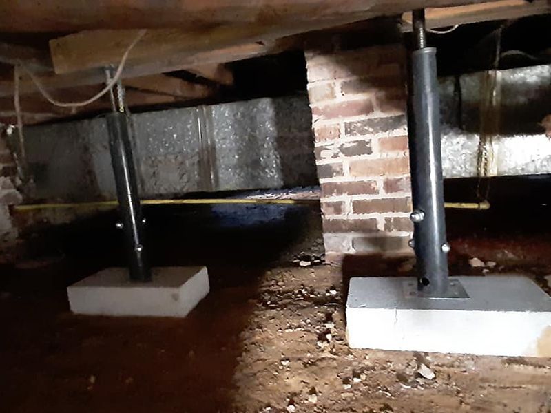 Two adjustable steel support jacks on concrete pads in a crawl space.