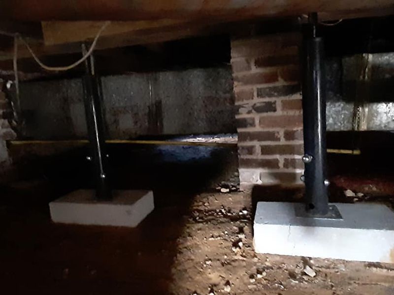 Two black adjustable columns support wooden beams in a dark, brick-walled crawl space, resting on gray concrete blocks.