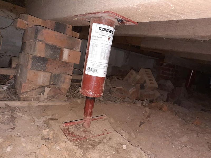 Red jack supporting a beam in a crawl space, resting on dirt and brick.