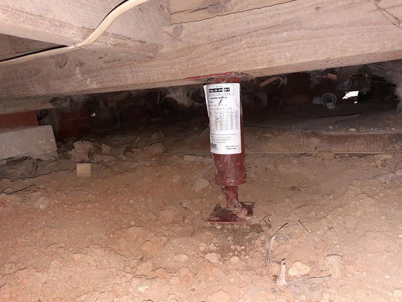 A red support jack beneath a wooden beam in a crawl space.