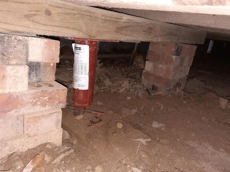 Red jack supporting a wooden beam in a crawl space with brick supports and dirt ground.