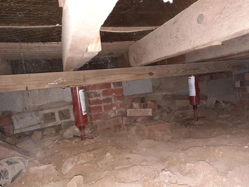 Underneath a house, a crawl space with red metal jacks supporting wooden beams over a dirt floor and brick structures.