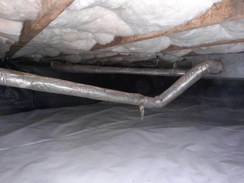 View inside a crawl space with insulated ducts, white plastic ground cover, and wooden beams.