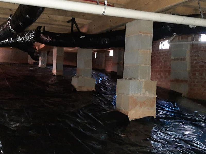 A crawl space with black plastic ground cover, concrete pillars, and ductwork.