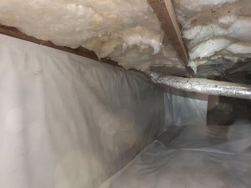 Crawl space with white vapor barrier on walls and floor, insulation above, and ductwork running across.