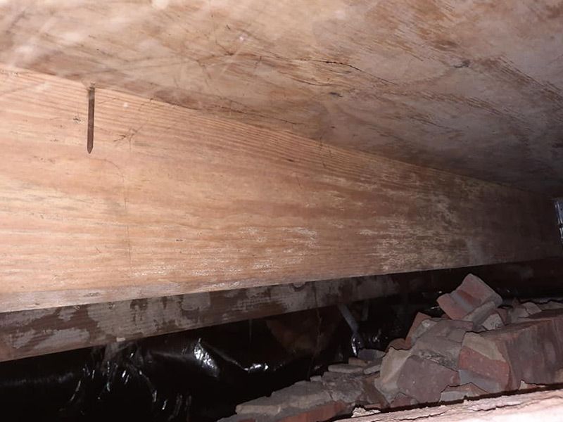 Wooden beam in a dark crawl space with brick rubble and black plastic sheeting visible below.