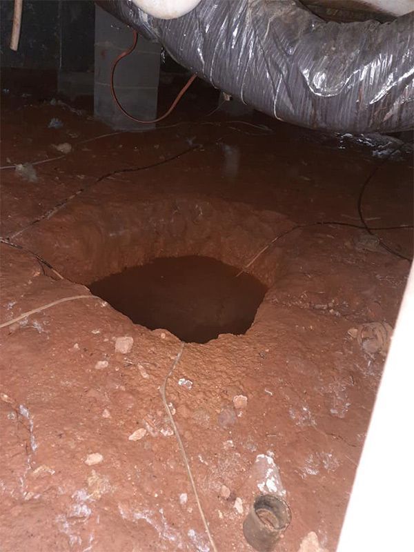Hole in the dirt floor of a crawlspace; reddish-brown soil, dark water at the bottom, air duct above.