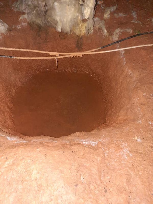 A deep, open, round hole filled with muddy water, dug into red soil under a white ceiling with wires.