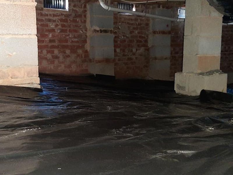 Black plastic sheeting covers a crawlspace floor with brick walls and concrete supports; water is present.