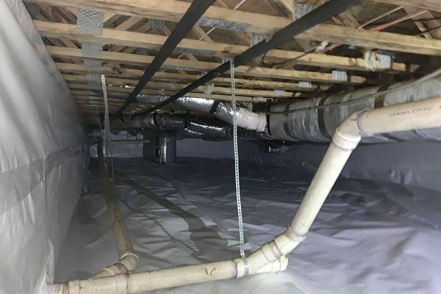 Crawl space with white vapor barrier on the floor and walls, ductwork, and pipes.