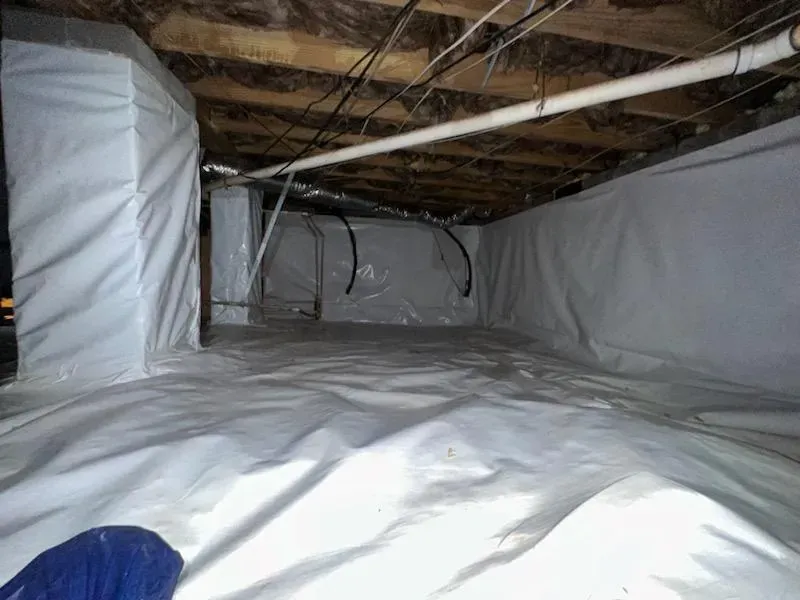 Crawl space covered in white vapor barrier. Wooden beams and pipes overhead.