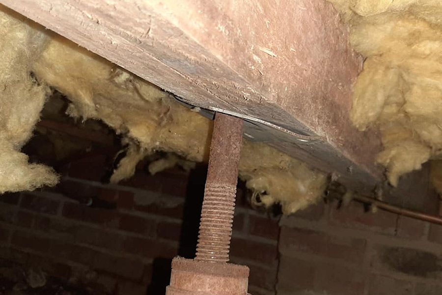 Rusty metal jack supporting a sagging wooden beam in an attic. Brown insulation and brick are visible.