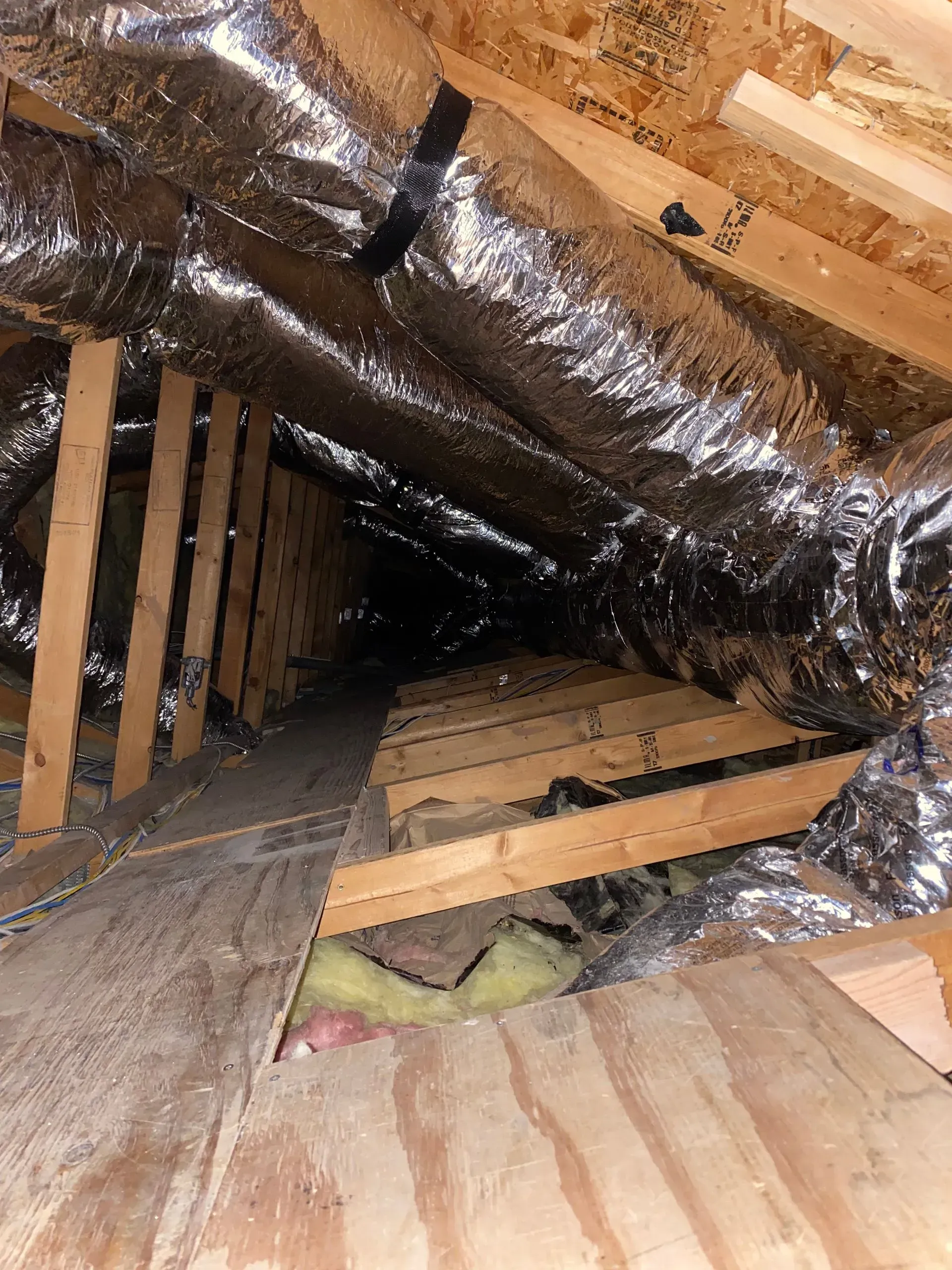 Insulated air ducts run through an attic space, positioned above wooden ceiling joists and plywood flooring.