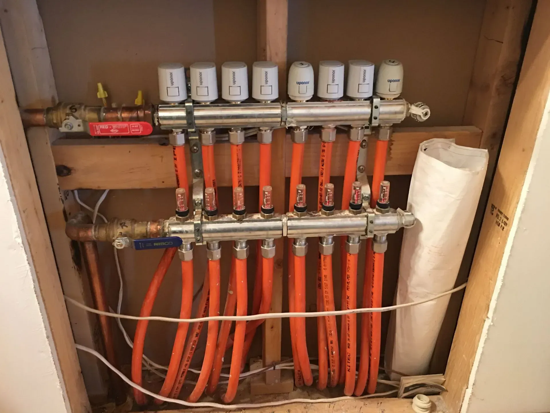 An interior view of a home heating system manifold with eight orange pipes connected to silver valves mounted on wood.