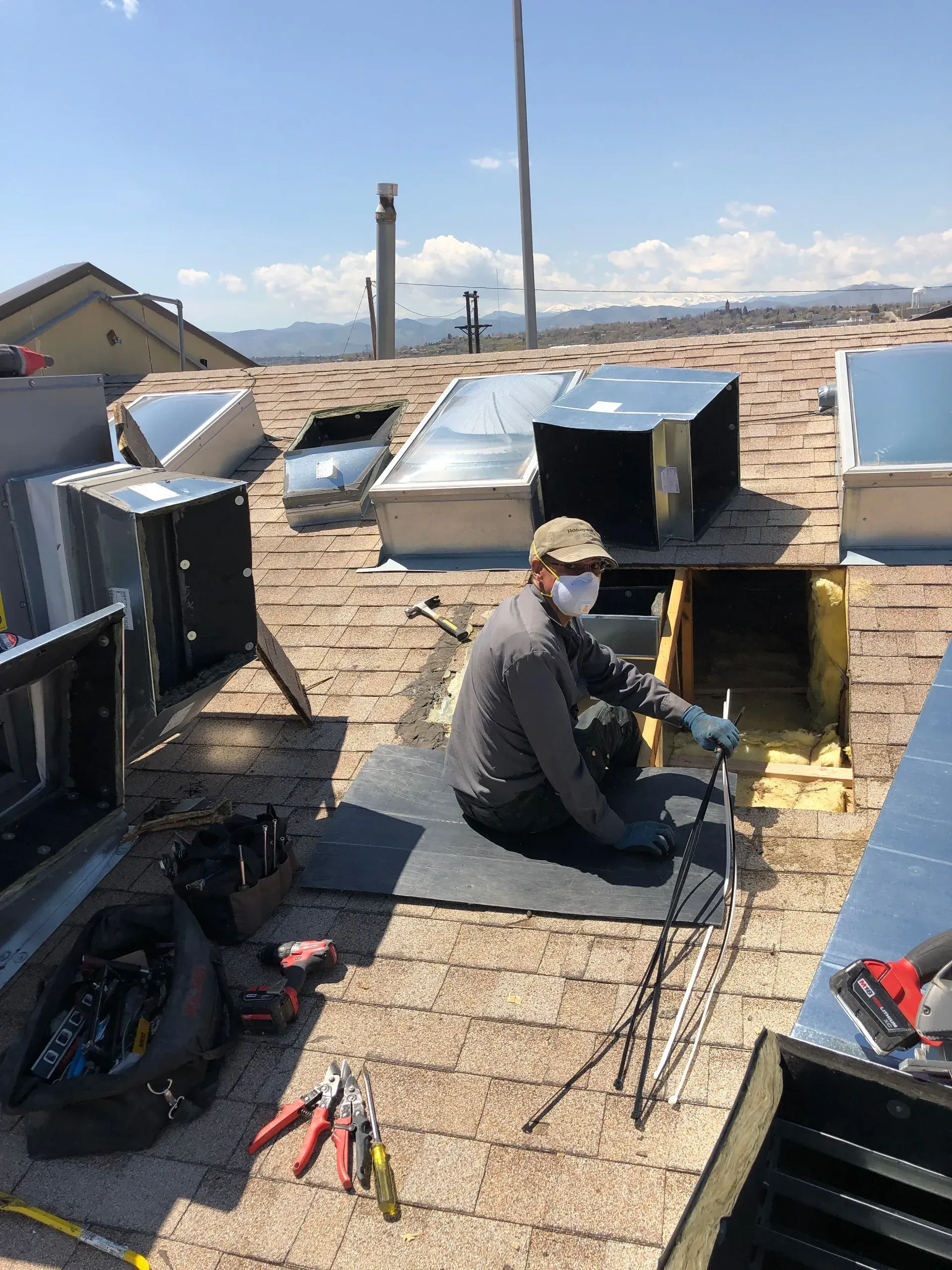 A technician wearing a mask and gloves works on an open residential roof, surrounded by tools and HVAC vent components.