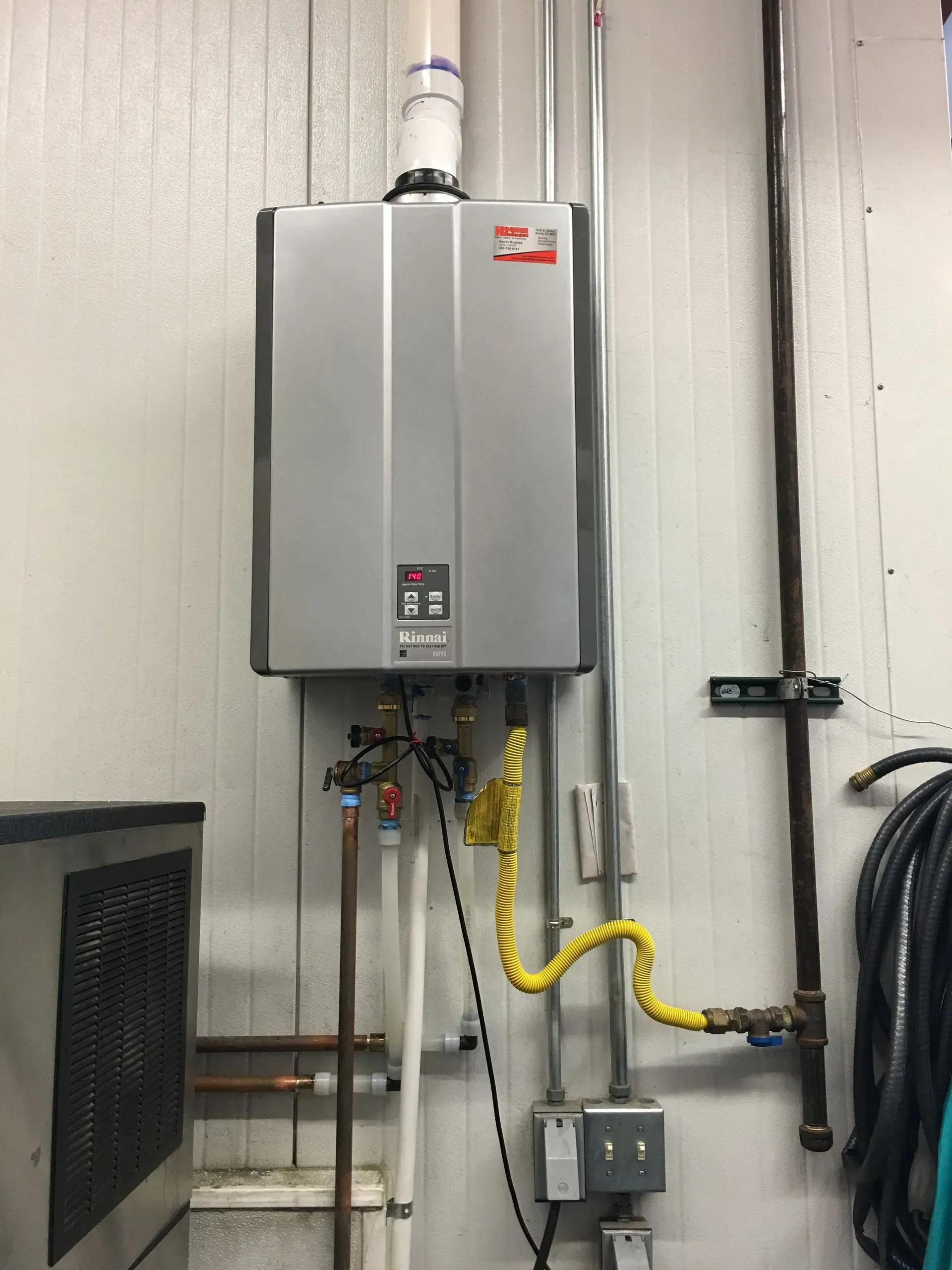 A wall-mounted Rinnai tankless water heater with gas and water supply lines in an industrial or commercial setting.