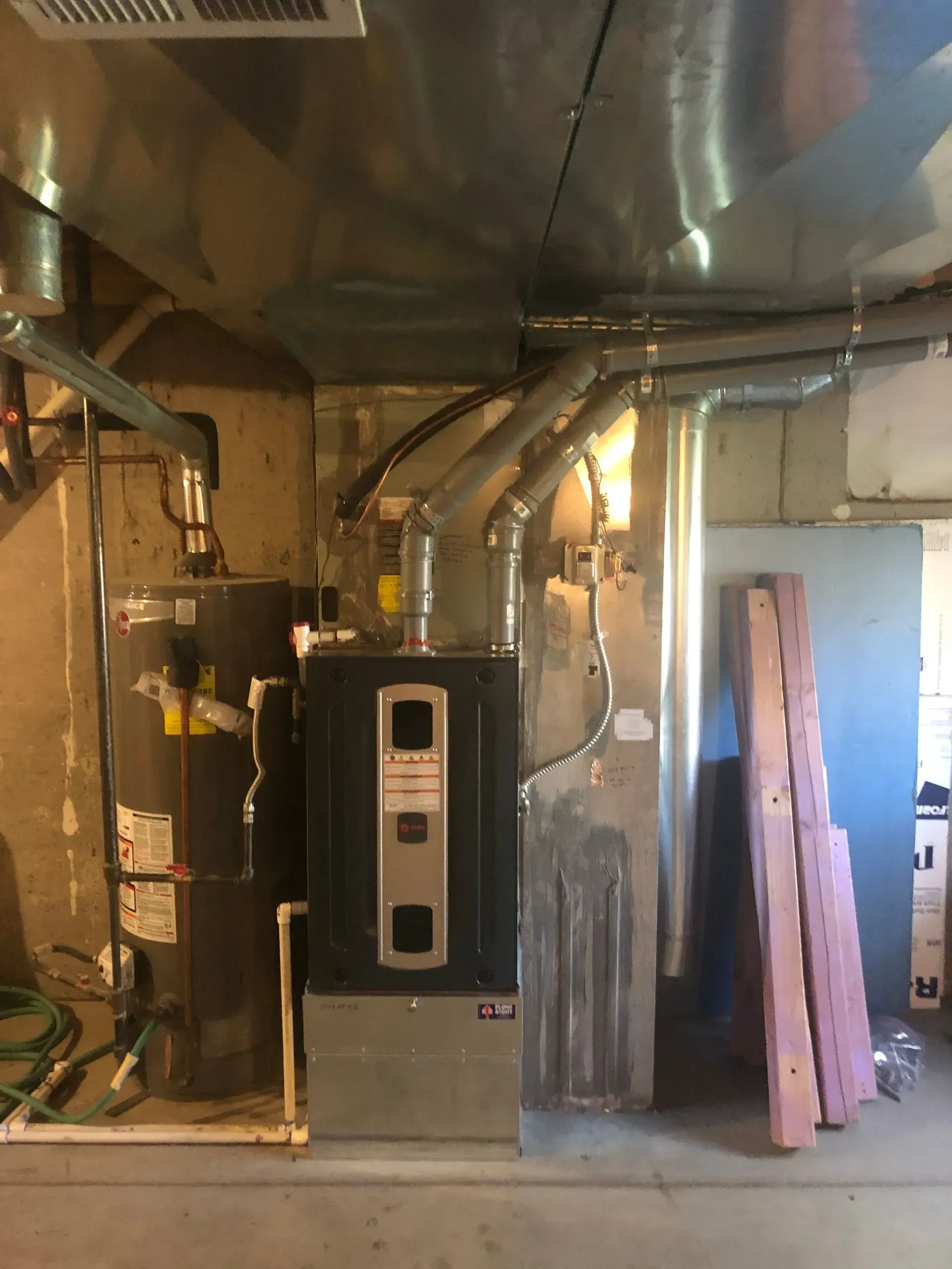 A water heater and a furnace unit stand next to each other in a basement with overhead metal ductwork.