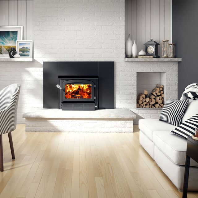 A lit fireplace insert centered on a white brick wall, with firewood in an alcove and a sofa in a bright living room.