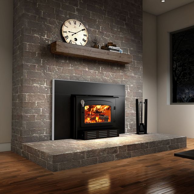A wood-burning stove insert glowing with a fire, set into a stone fireplace with a wooden mantel and clock.