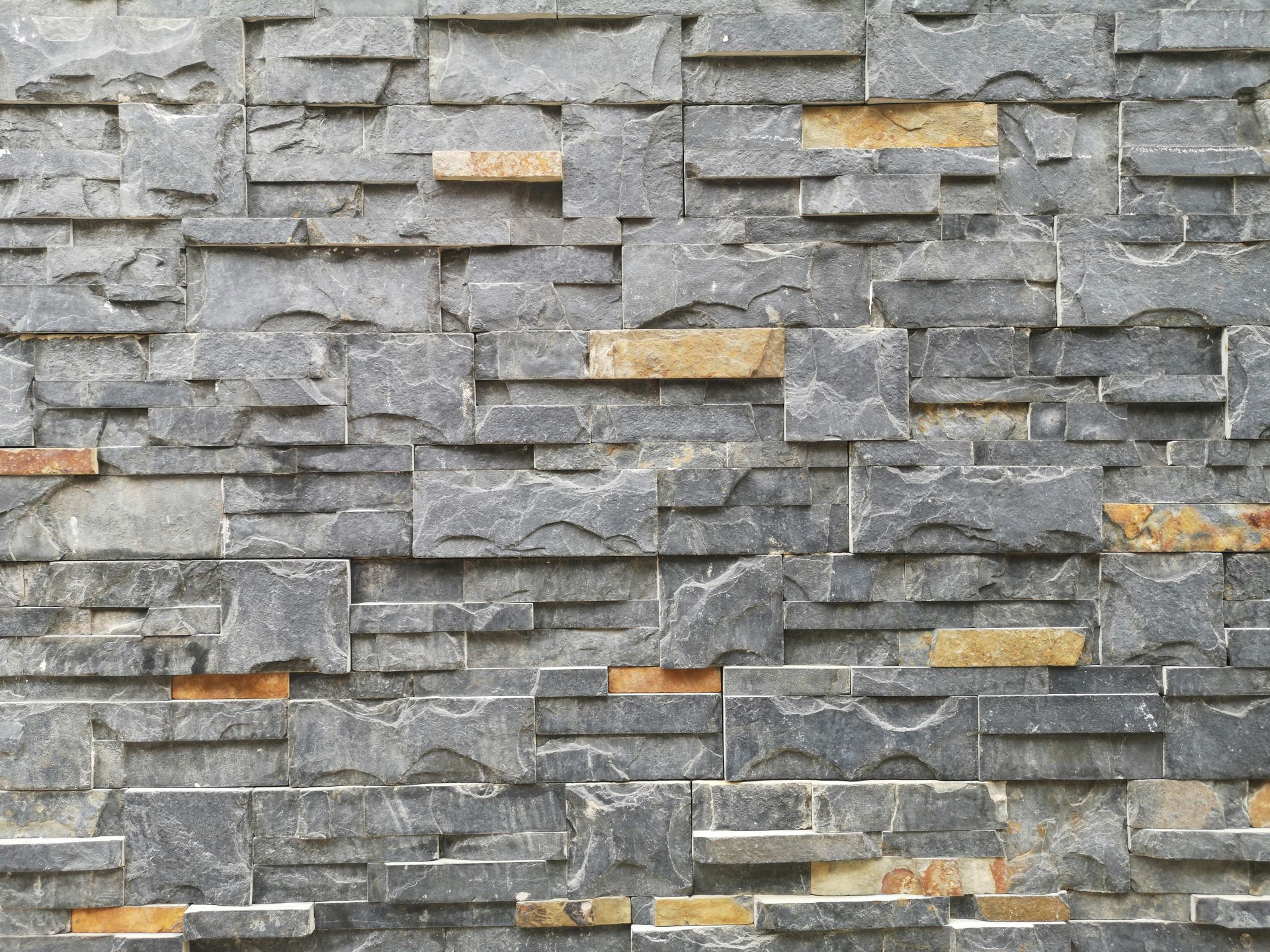 A textured wall made of stacked grey slate bricks with scattered accents of tan and golden stone.