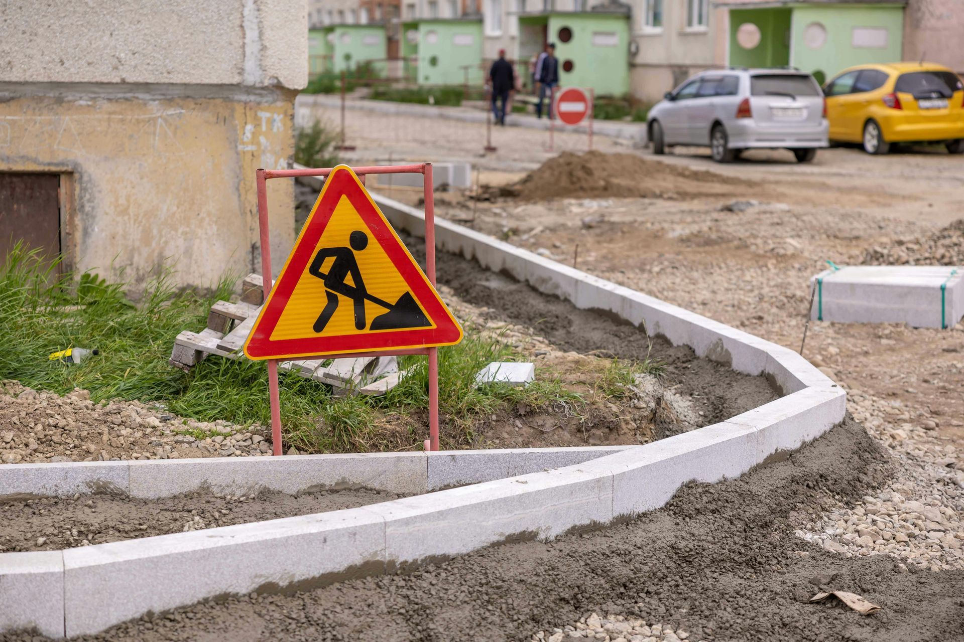 A yellow and red construction sign stands near a new curb under installation in a residential neighborhood.