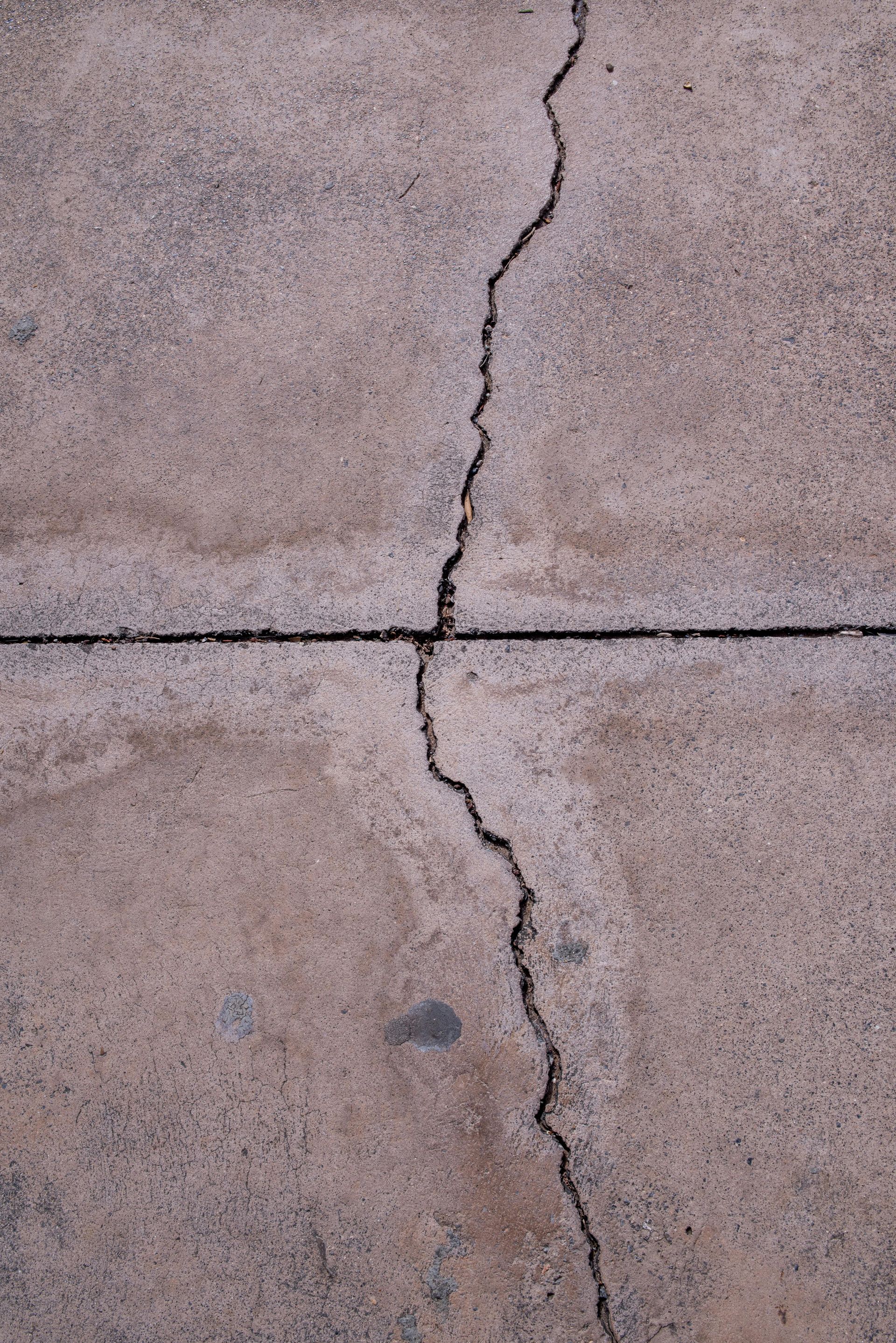 A vertical jagged crack runs through a concrete slab divided by a straight horizontal control joint.