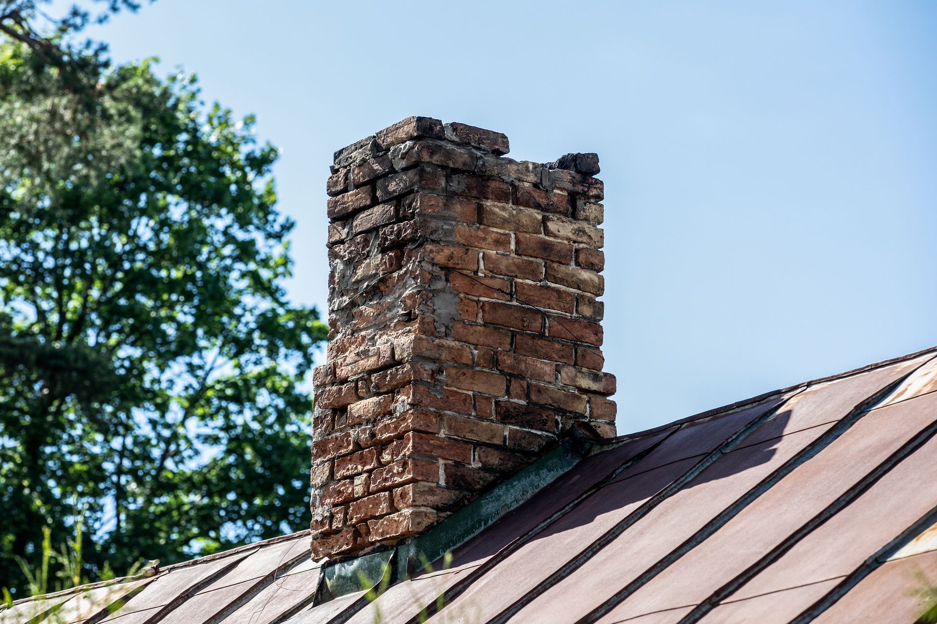 Brick chimney on a metal roof, with a clear blue sky and trees in the background.
