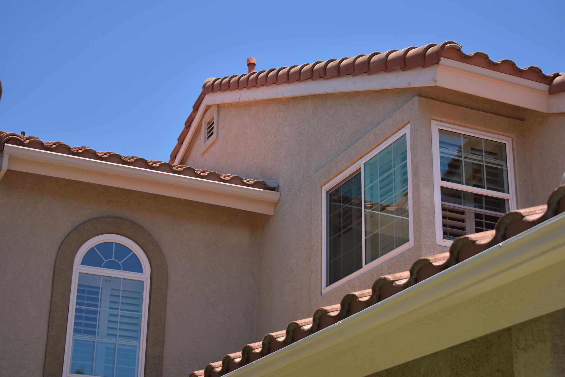 Exterior view of a beige house with a tan stucco facade, a red tiled roof, and several large windows under a blue sky.