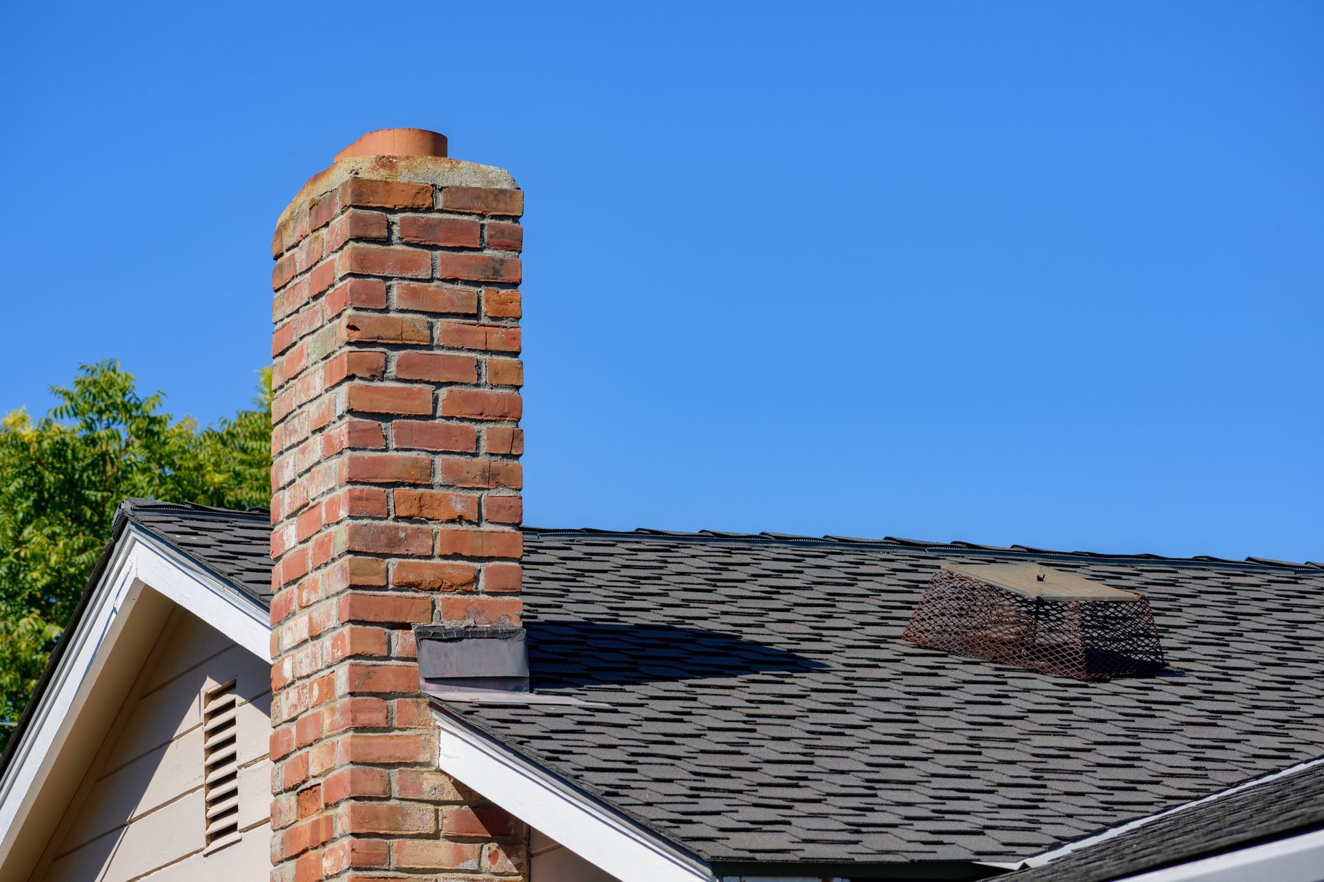 A brick chimney rises from a shingled roof against a clear blue sky, next to a small attic vent.