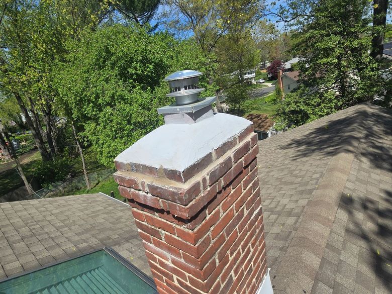 Brick chimney on a roof with a metal cap, trees in the background.