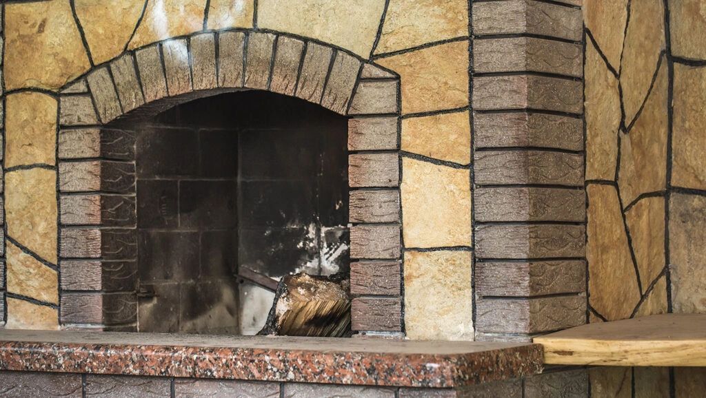 Stone fireplace with arched opening, dark interior, brown and tan stone exterior, and a wood shelf.