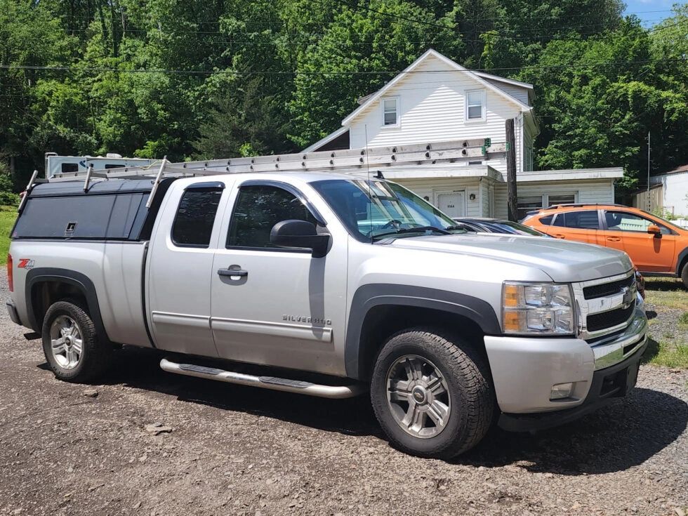 Silver Chevy pickup truck with ladder rack parked outdoors near a house and trees.