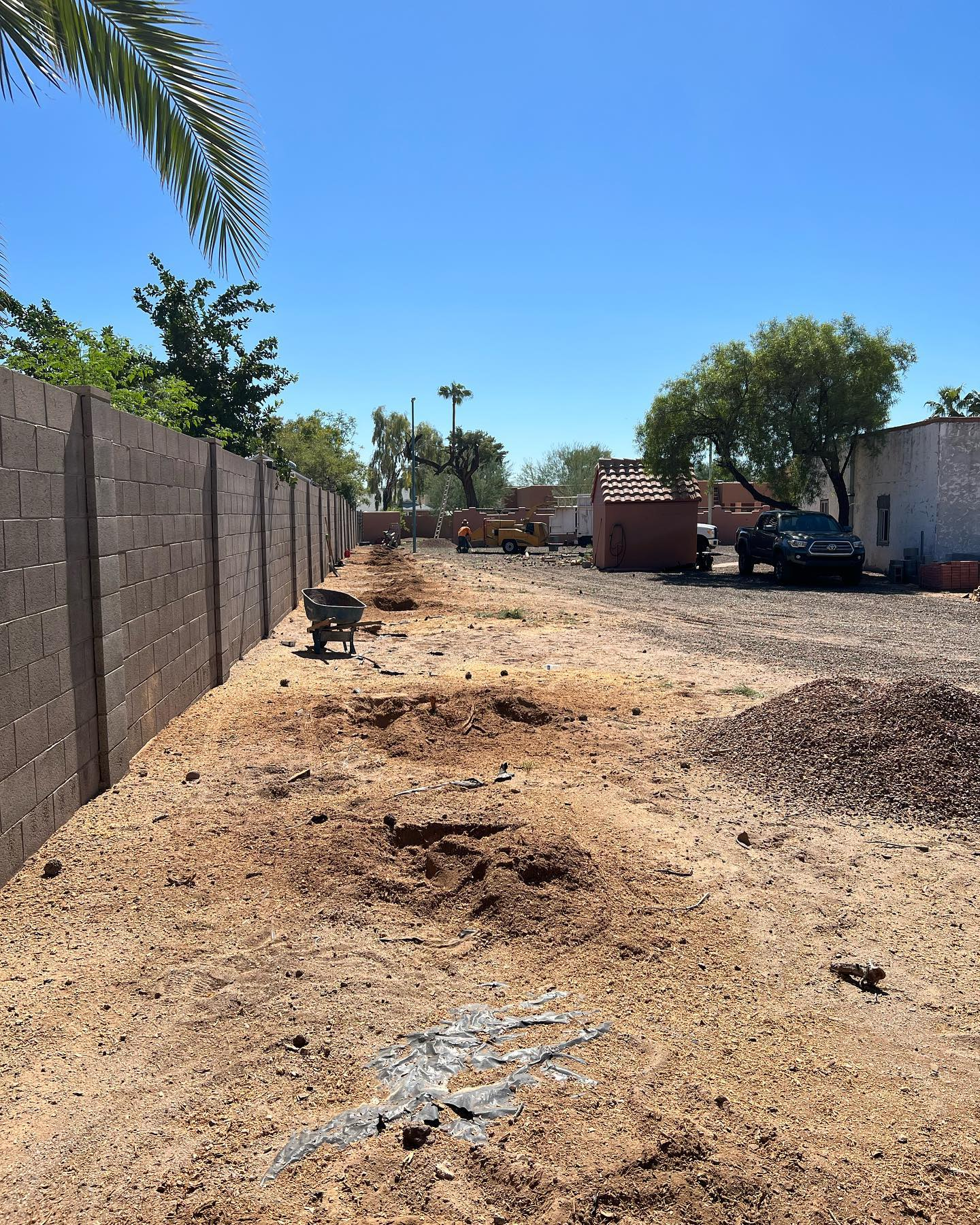 A brick fence surrounds a dirt field with a wheelbarrow in the foreground